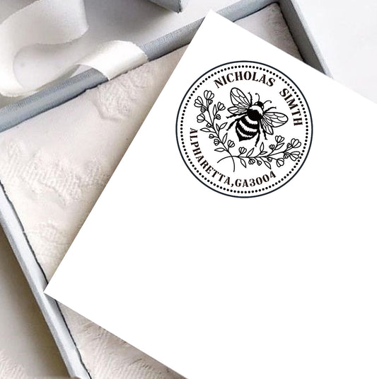 A personalized self inking address stamp, customized with your name and address, bee and flower, stamped on the white card, a gift box is under it.
