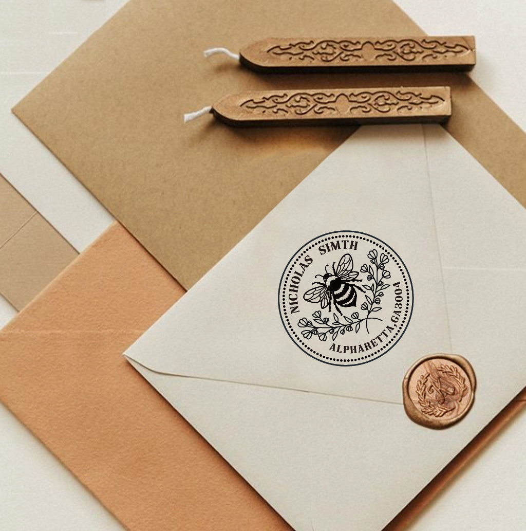 A personalized self inking return address stamp, customized with your name and address, bee and flower, stamped on the white envelope, beside it, sealing wax is creating a wax seal on the mail.