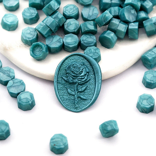 Some pearlescent dark teal wax beads placed on white cushion and cloth, and an wax seal created with them.