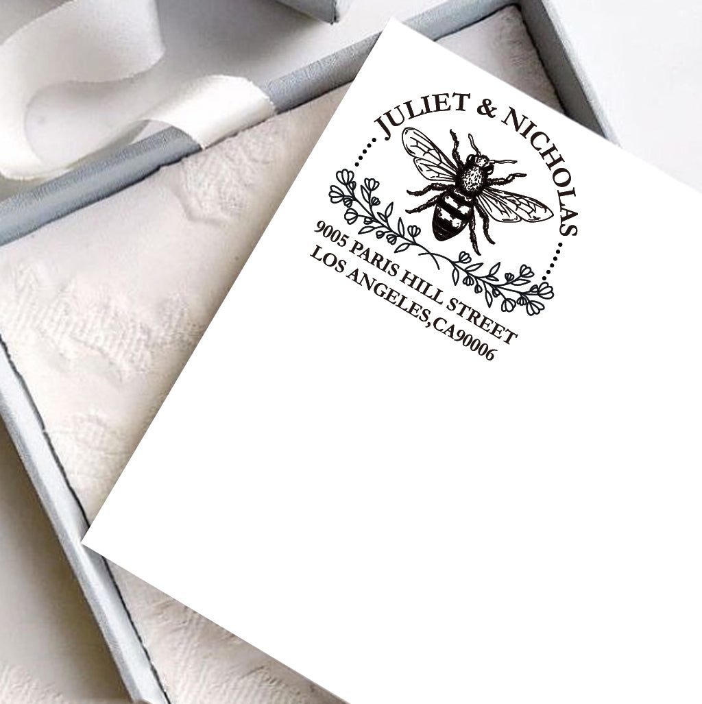A personalized self inking address stamp, customized with your name and address, bee and flower, stamped on the white card, a gift box is under it.
