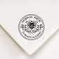 An address stamp's artwork on the white envelope.Inside a circle, 2 staggered floral branch under the bee at the middle.The custom name above the bee and the address under the floral,they are made in curve.