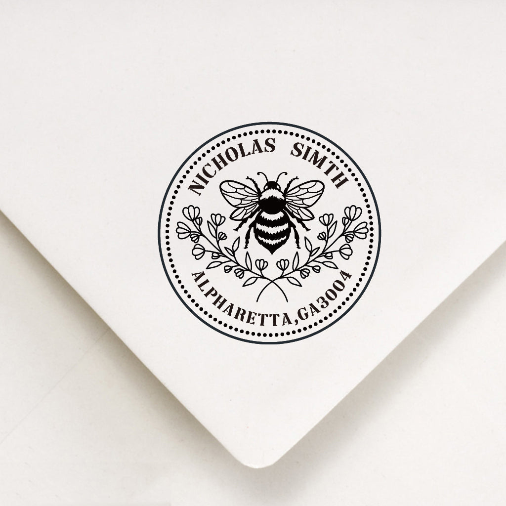 An address stamp's artwork on the white envelope.Inside a circle, 2 staggered floral branch under the bee at the middle.The custom name above the bee and the address under the floral,they are made in curve.