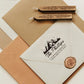 A personalized self inking return address stamp, customized with your mountain name and address, stamped on the white envelope, beside it, sealing wax is creating a wax seal on the mail.