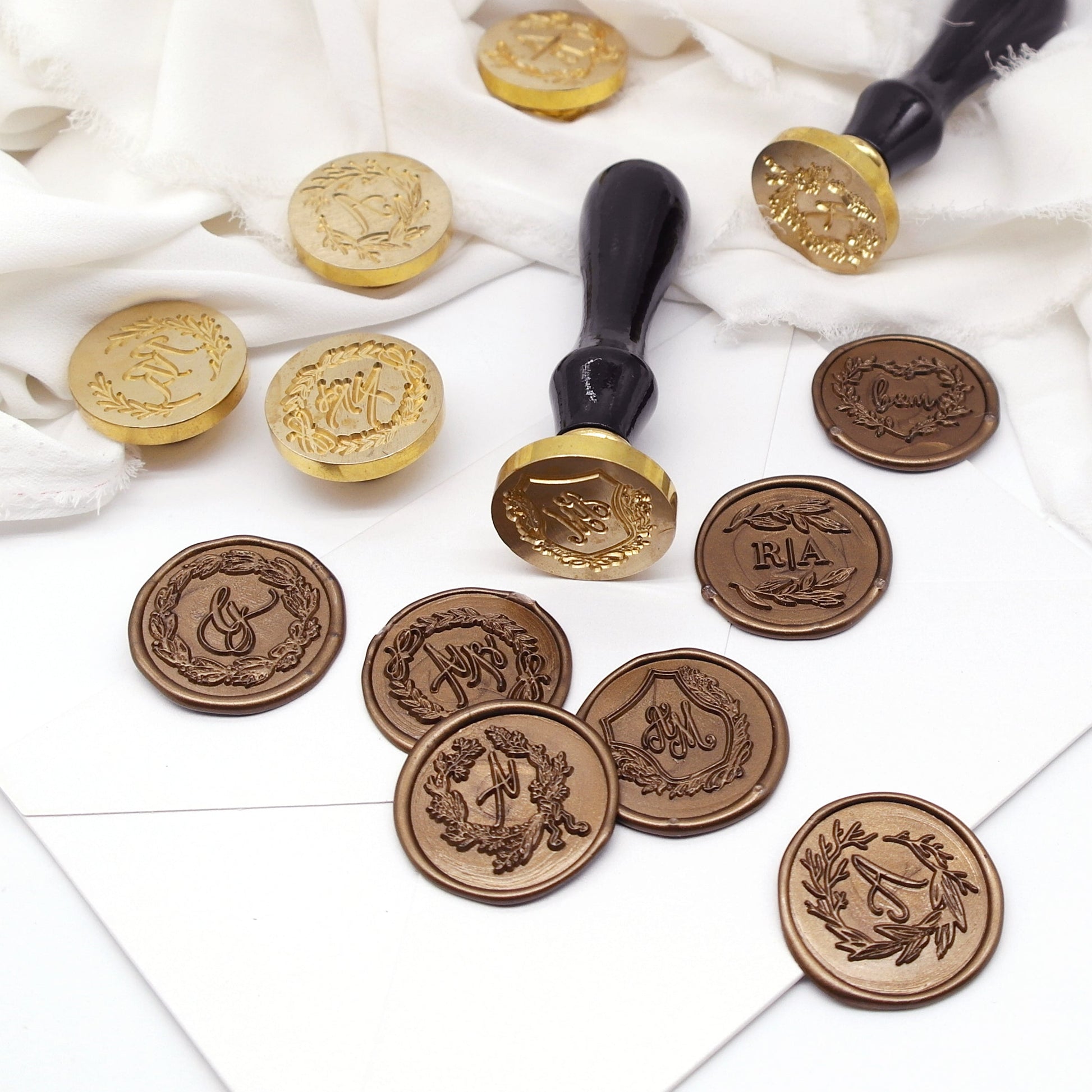 Seven wedding wax seals with wreath and monogram artwork created on a white envelope.Two wax stamps with black wooden handle lie down on the envelope and white cloth,nearby,standing four brass heads.