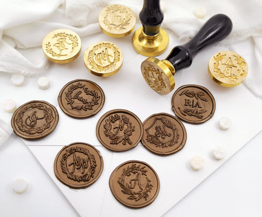 Seven wedding wax seals with wreath and monogram artwork created on a white envelope.One wax stamp with black wooden handle lie down on the corner of the envelope and white cloth,nearby,standing a wax stamp and four brass heads. And white octagon wax beads surrounding.